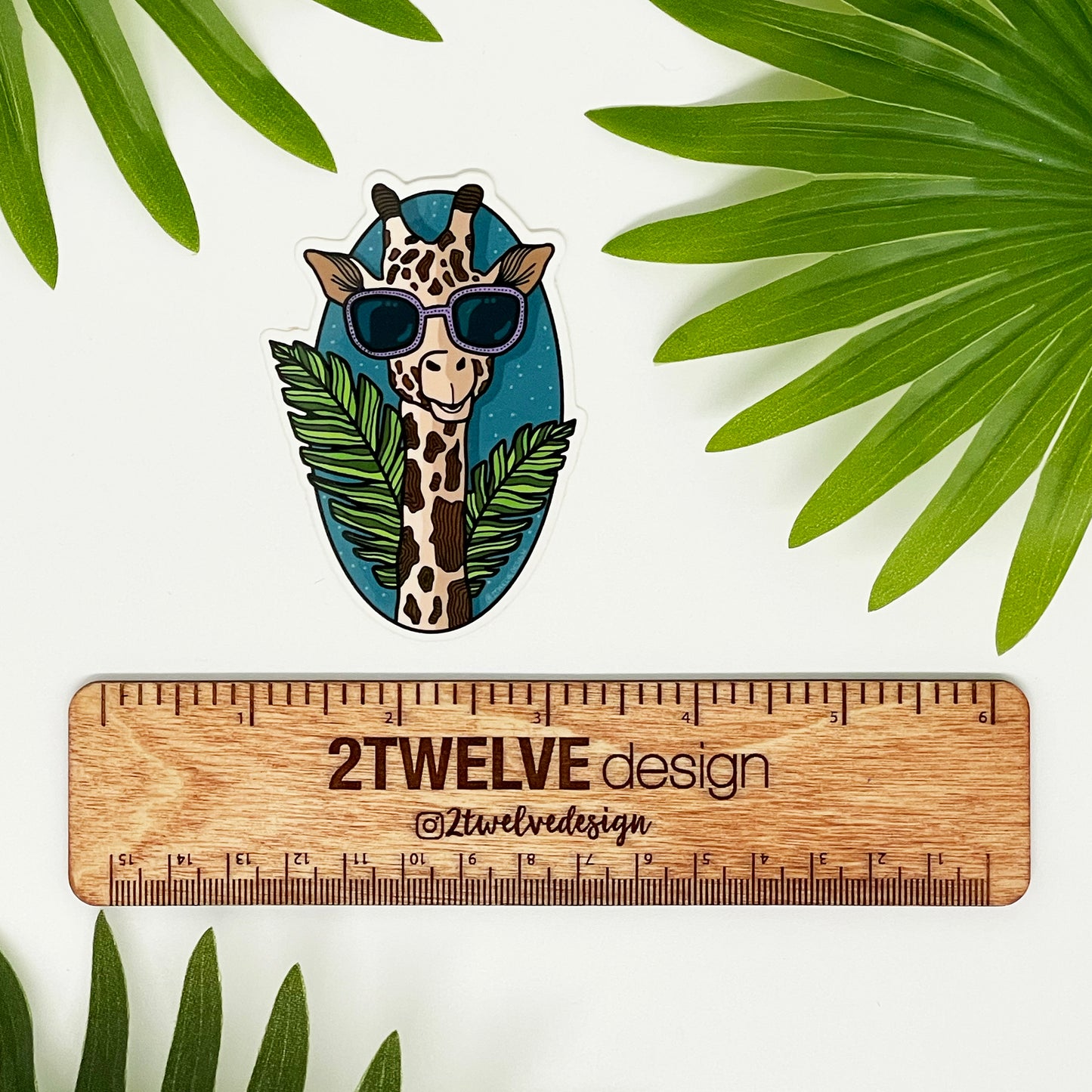 A quirky illustration of a giraffe made into a durable vinyl sticker laying on top of a white background surrounded by leaves and a ruler to show actual size.