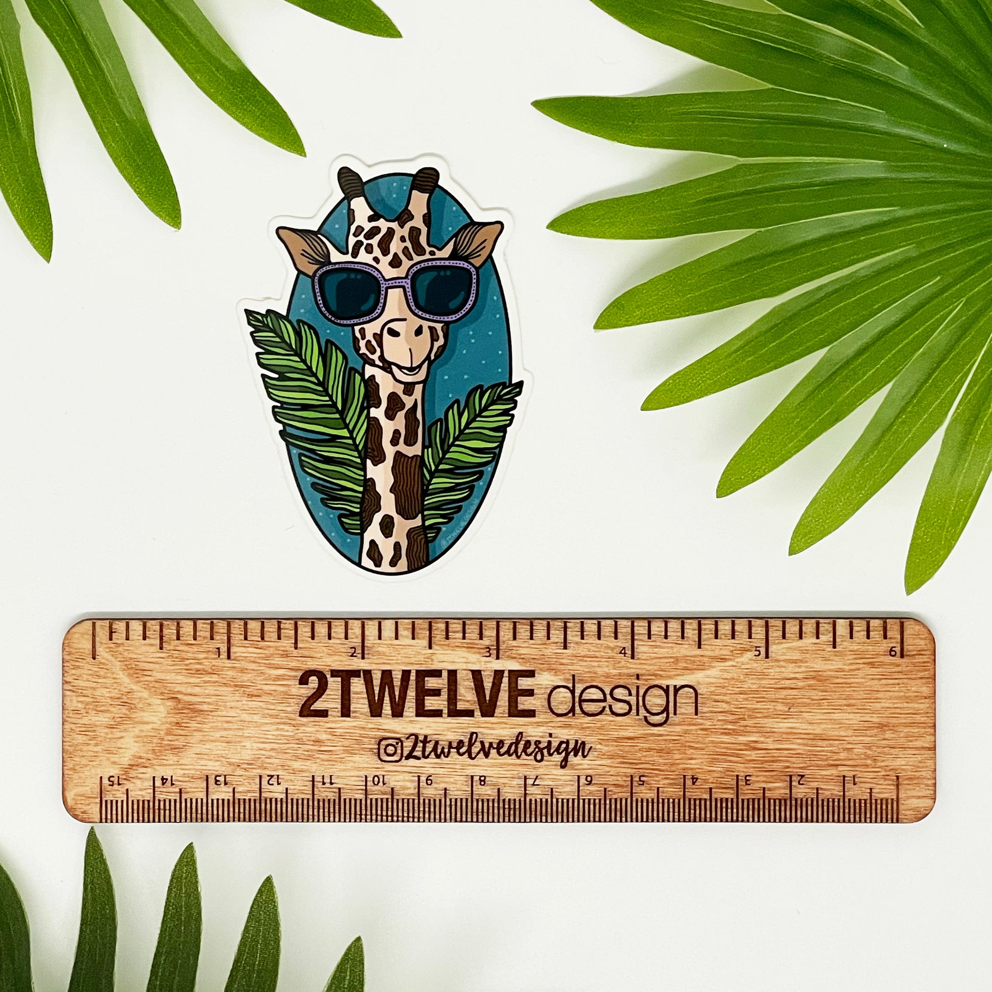 A quirky illustration of a giraffe made into a durable vinyl sticker laying on top of a white background surrounded by leaves and a ruler to show actual size.