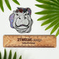 A hippo illustration made into a glittery diecut sticker. Laying on a white background surrounded by palms. "2Twelve Design" ruler shows actual size