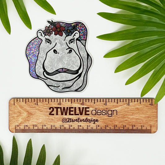 A hippo illustration made into a glittery diecut sticker. Laying on a white background surrounded by palms. "2Twelve Design" ruler shows actual size