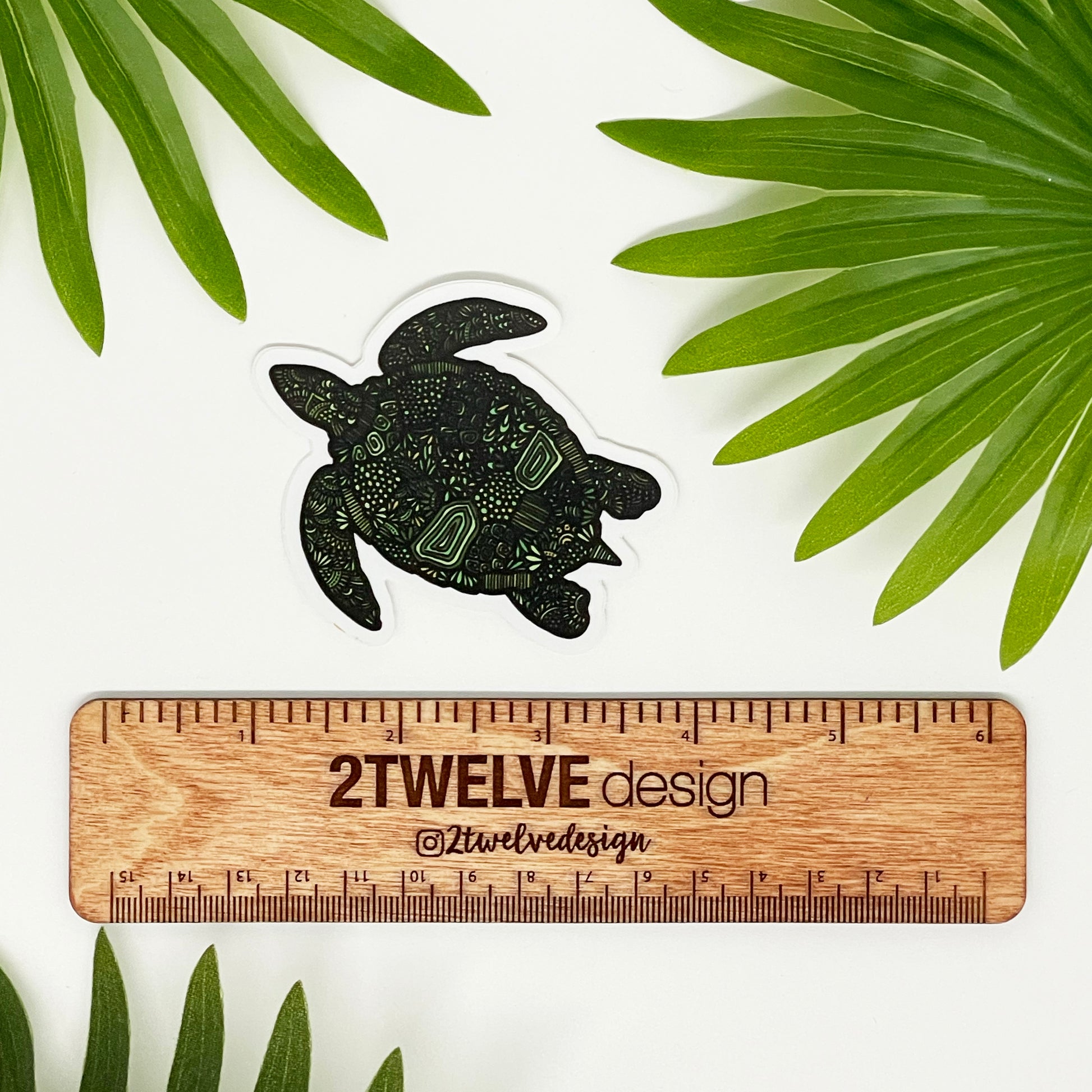 A green turtle sticker laying on a white surface above a ruler that says "2Twelve Design" surrounded by palm leaves
