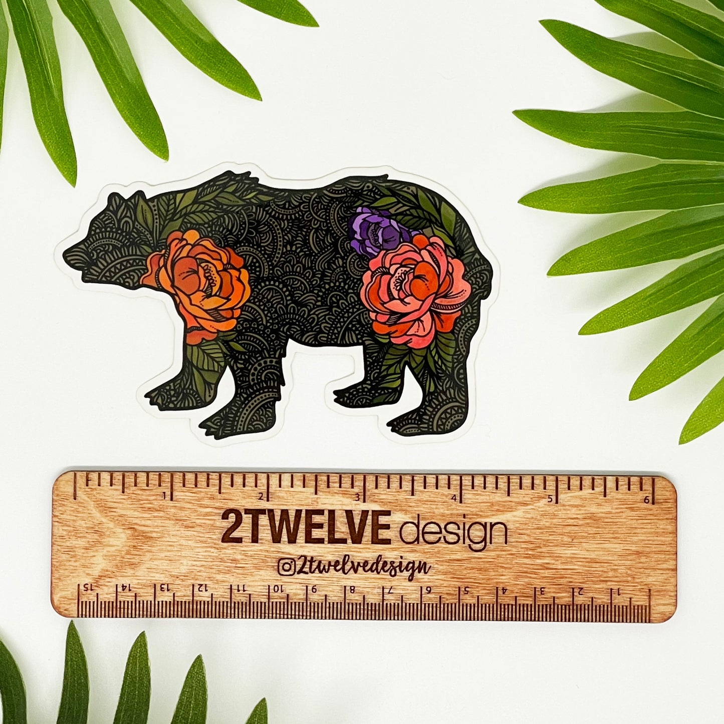 An intricate illustrated bear made into a vinyl sticker. Laying on a white surface above a "2Twelve Design" wooden ruler. Surrounded by palm leaves.