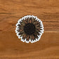A very intricate sunflower drawing made into a die-cut matte sticker. Laying on a wooden surface.