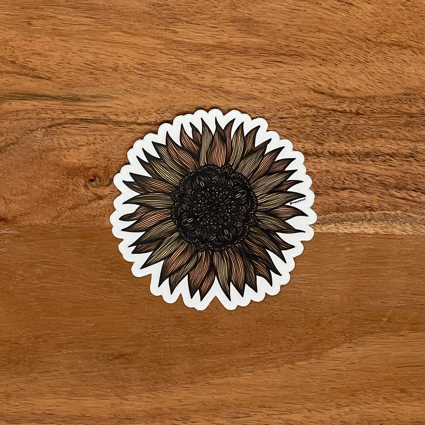 A very intricate sunflower drawing made into a die-cut matte sticker. Laying on a wooden surface.