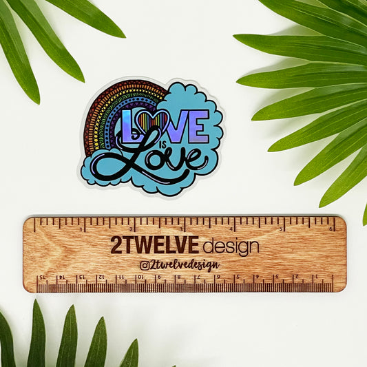 A hand drawn illustration of "Love is Love" with a rainbow and clouds, turned into a die-cut sticker. Laying on a white background above a "2Twelve Design" ruler. Surrounded by palm leaves.