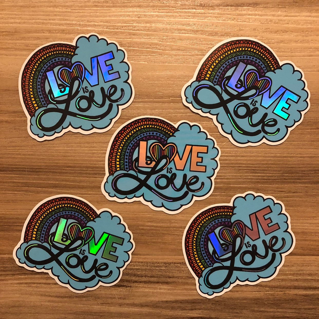 A group of Love is Love rainbow holographic stickers laying on a wooden background.