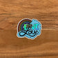 A die-cut holographic sticker with a hand-drawn love is love illustration, featuring a rainbow and the phrase 'love is love' in a circular blue font.