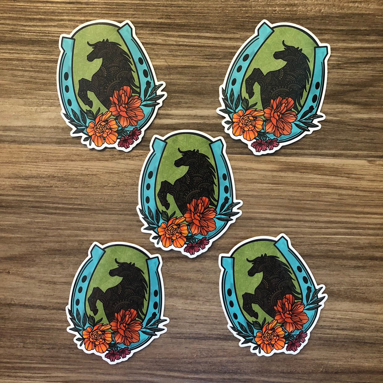A hand-drawn horse and horseshoe illustration made into a vinyl die-cut sticker. A group of five stickers laying on a wooden surface.