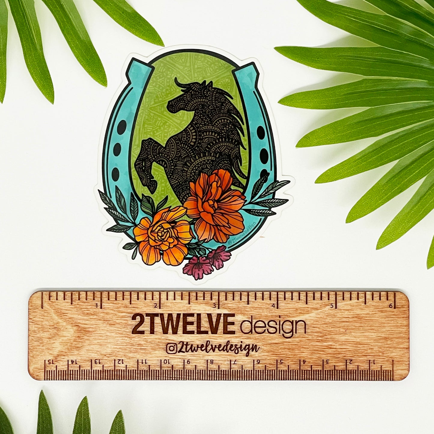 A vinyl, waterproof die-cut sticker featuring a hand-drawn floral and horse illustration, with a horseshoe shape at the top, placed on a wooden surface with green foliage in the background.