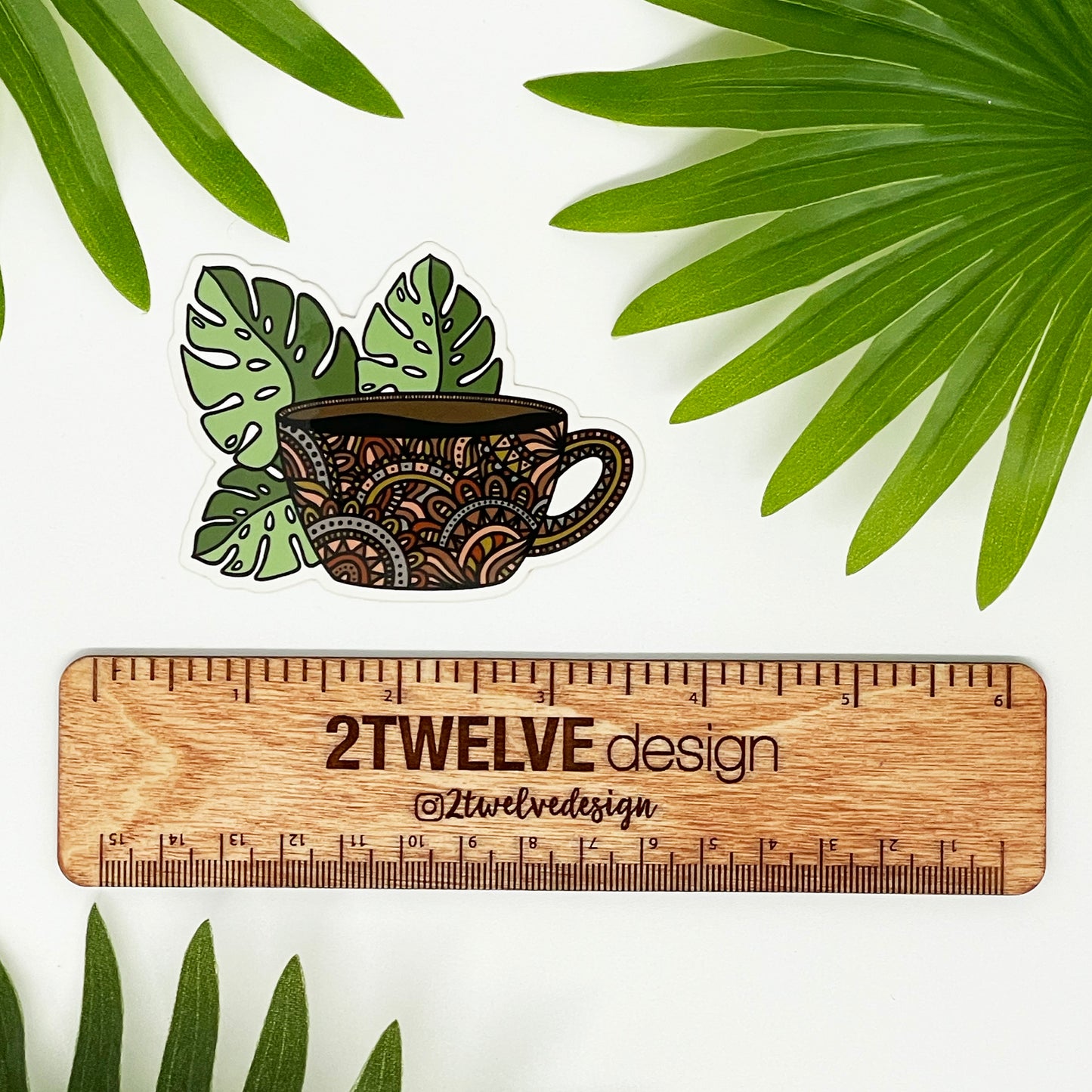 A die-cut vinyl sticker featuring a hand-drawn illustration of a coffee mug with monstera leaves laying on a white background above a "2Twelve Design" ruler, surrounded by palm leaves.