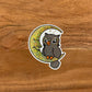 A cute and fun owl illustration made into a die-cut, glitter sticker. Laying on a wooden surface.