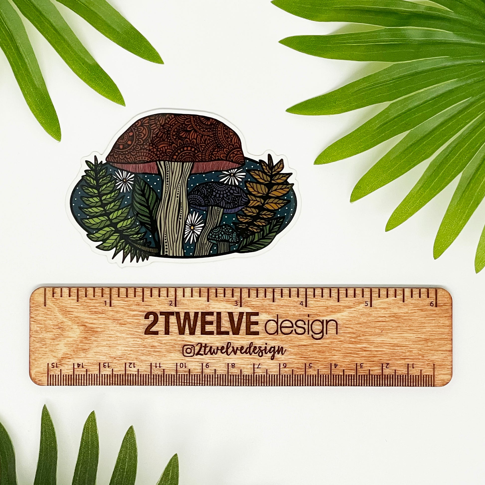 A woodland themed mushroom diecut sticker laying on a white background above a "2Twelve Design" ruler. Surrounded by palm leaves.