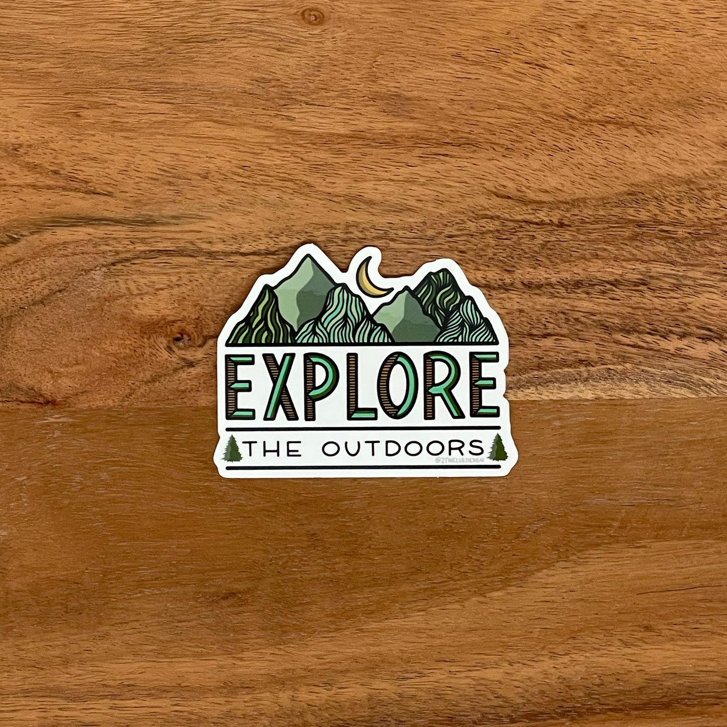 Mountain and moon illustration with "Explore The Outdoors" made into a vinyl sticker. Laying on a wooden surface.