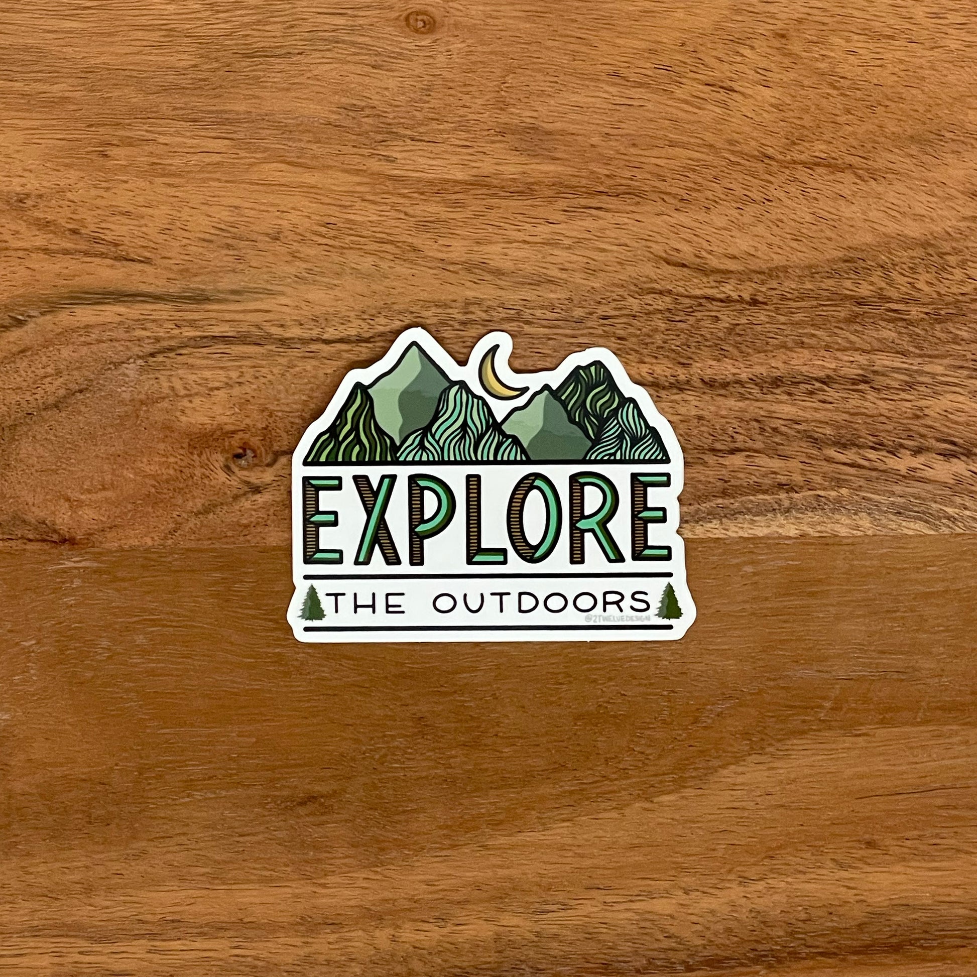 Mountain and moon illustration with "Explore The Outdoors" made into a vinyl sticker. Laying on a wooden surface.