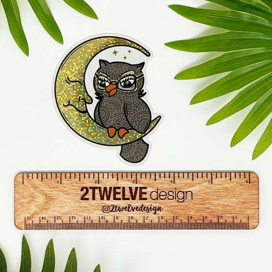 A cute, detailed owl and moon illustration made into a vinyl glitter die-cut sticker. Laying on a white surface above a "2Twelve Design" ruler to show size.