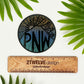 A "PNW" mountain illustration made into a vinyl circle sticker. Laying on a white surface above a "2Twelve Design" ruler. Surrounded by palm leaves.