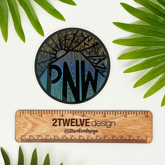 A "PNW" mountain illustration made into a vinyl circle sticker. Laying on a white surface above a "2Twelve Design" ruler. Surrounded by palm leaves.