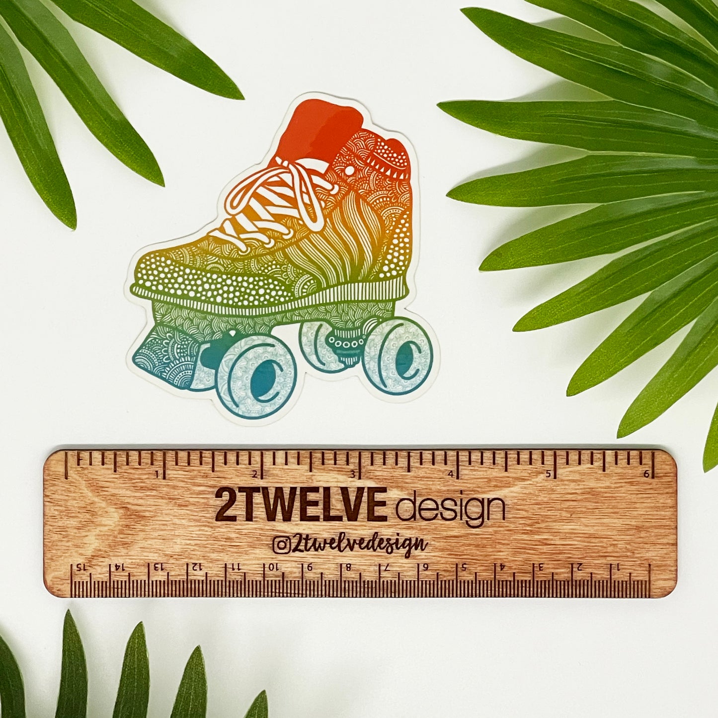 A rainbow color illustration of a roller skate turned into a die-cut sticker. Laying on a white surface surrounded by green palm leaves.