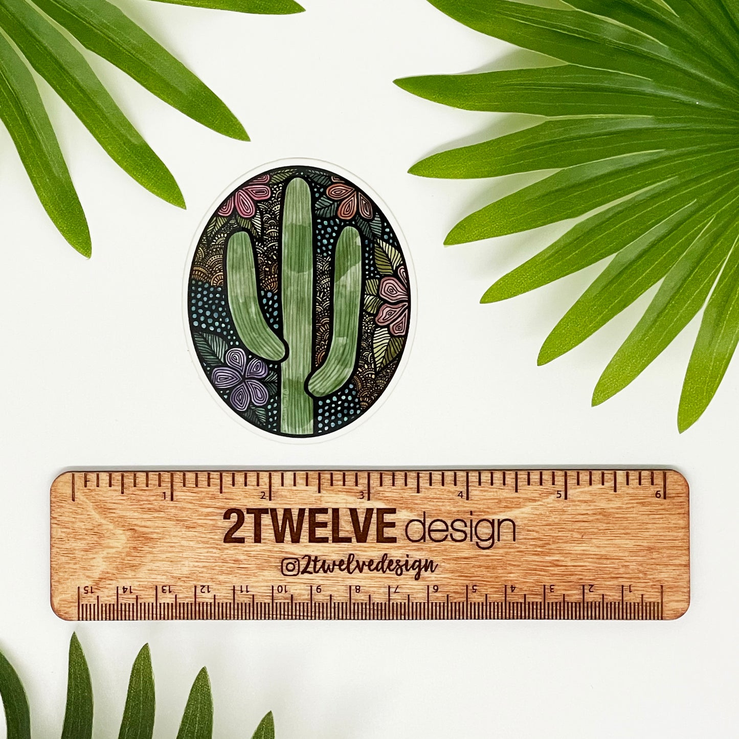 A floral and cactus oval sticker. Pastel colors and cute details. Laying above a "2Twelve Design" ruler on a white background surrounded by palm leaves. 