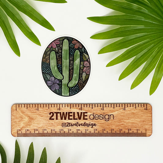 A floral and cactus oval sticker. Pastel colors and cute details. Laying above a "2Twelve Design" ruler on a white background surrounded by palm leaves. 