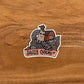 A sassy chicken illustration made into a vinyl die-cut sticker with a "Sassy Chick" banner. Laying on a wood surface.