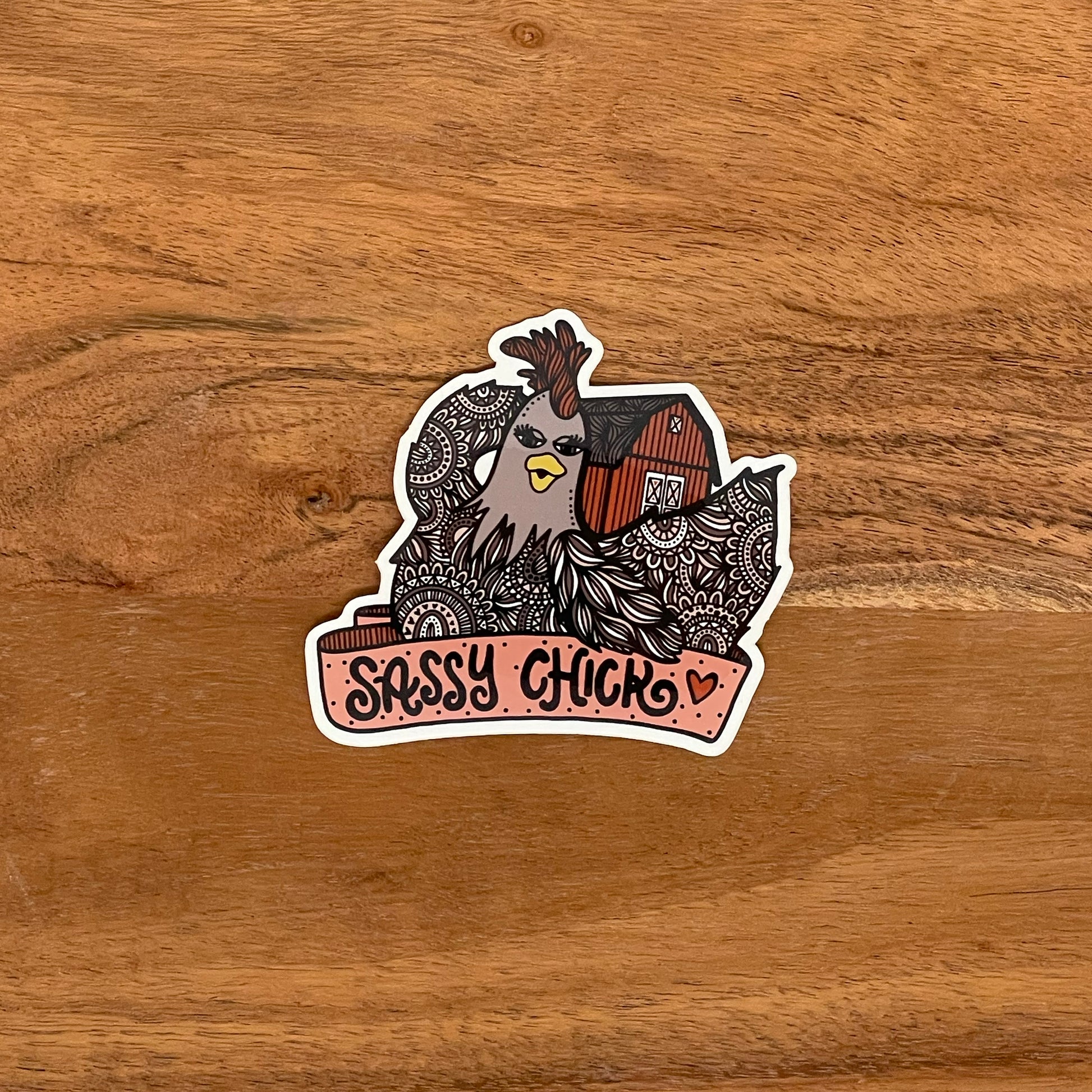 A sassy chicken illustration made into a vinyl die-cut sticker with a "Sassy Chick" banner. Laying on a wood surface.