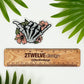 A vinyl sticker featuring a hand-drawn illustration of a skeleton shaka with floral elements and a matte shimmer finish, placed on a diecut sticker holder.