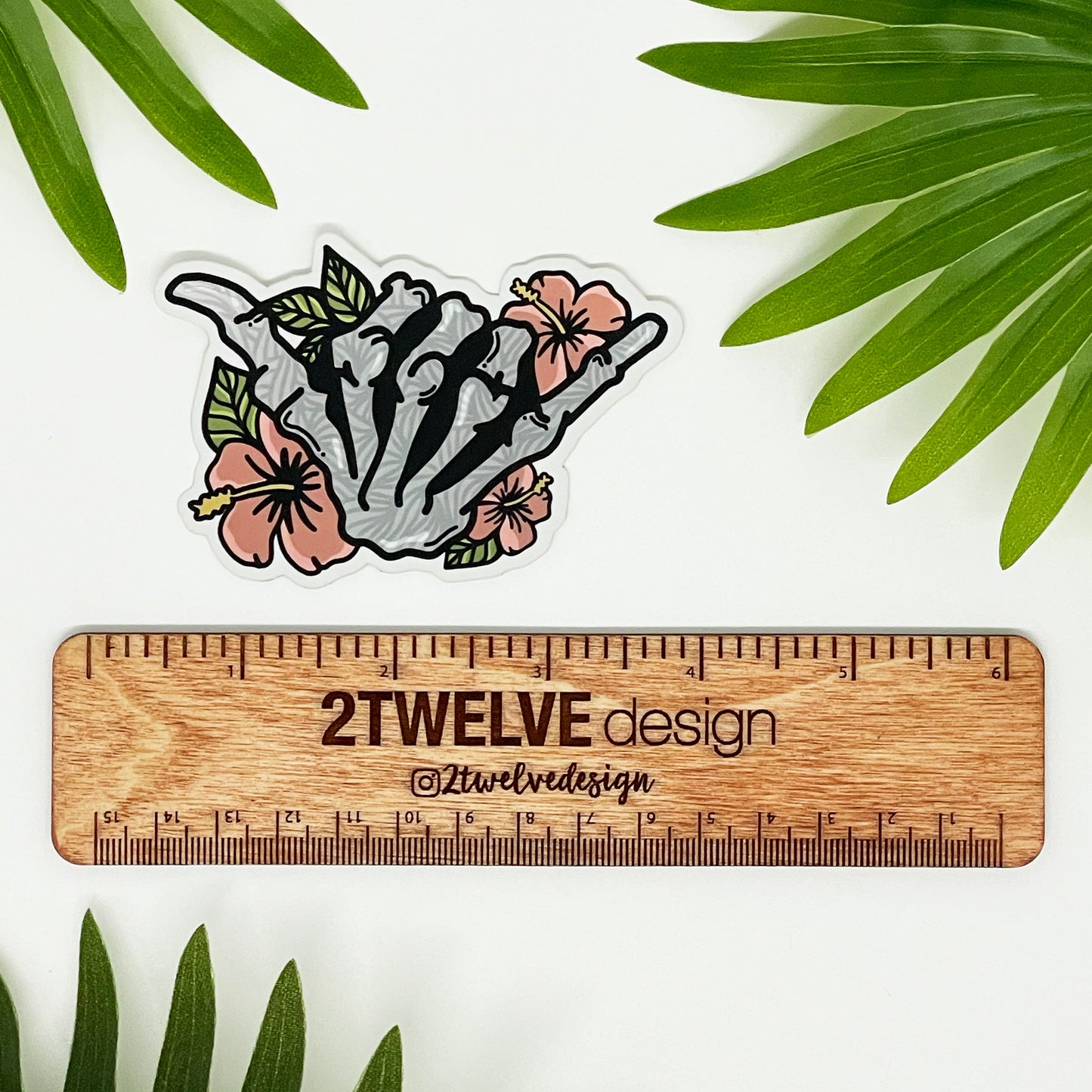 A vinyl sticker featuring a hand-drawn illustration of a skeleton shaka with floral elements and a matte shimmer finish, placed on a diecut sticker holder.