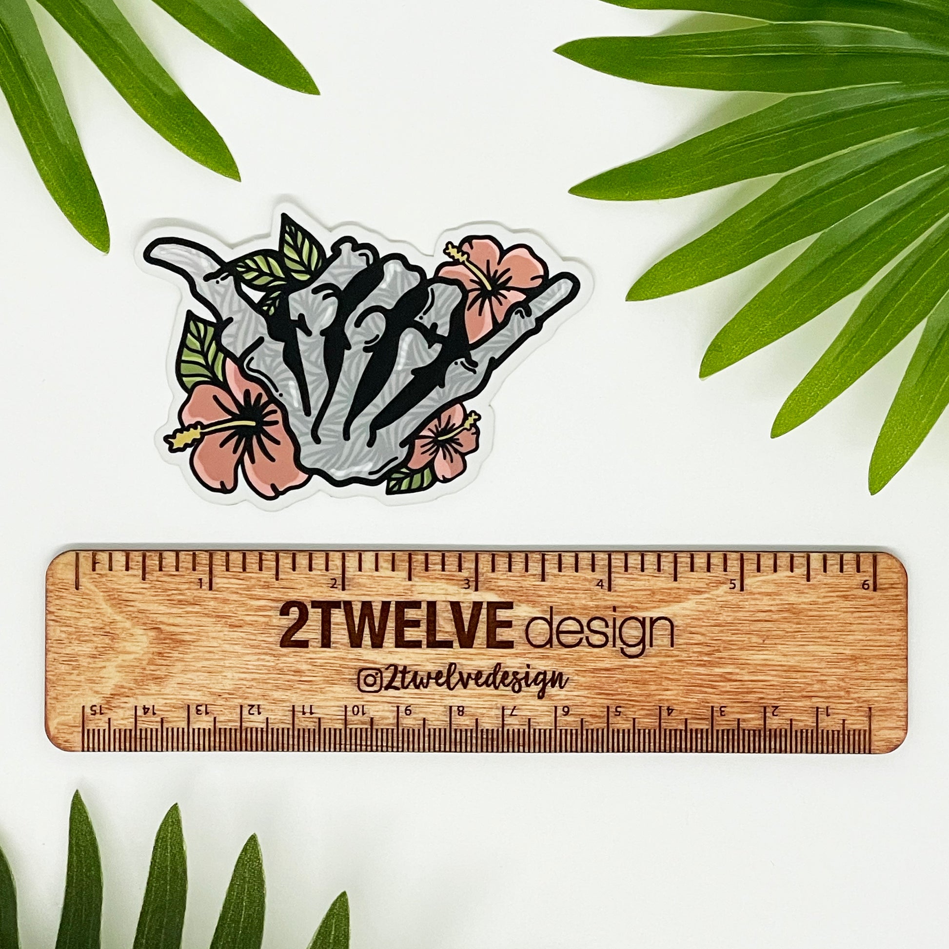 A vinyl sticker featuring a hand-drawn illustration of a skeleton shaka with floral elements and a matte shimmer finish, placed on a diecut sticker holder.