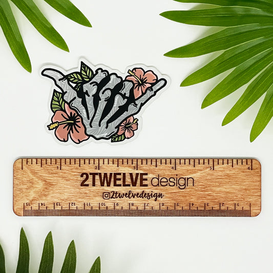 A vinyl sticker featuring a hand-drawn illustration of a skeleton shaka with floral elements and a matte shimmer finish, placed on a diecut sticker holder.