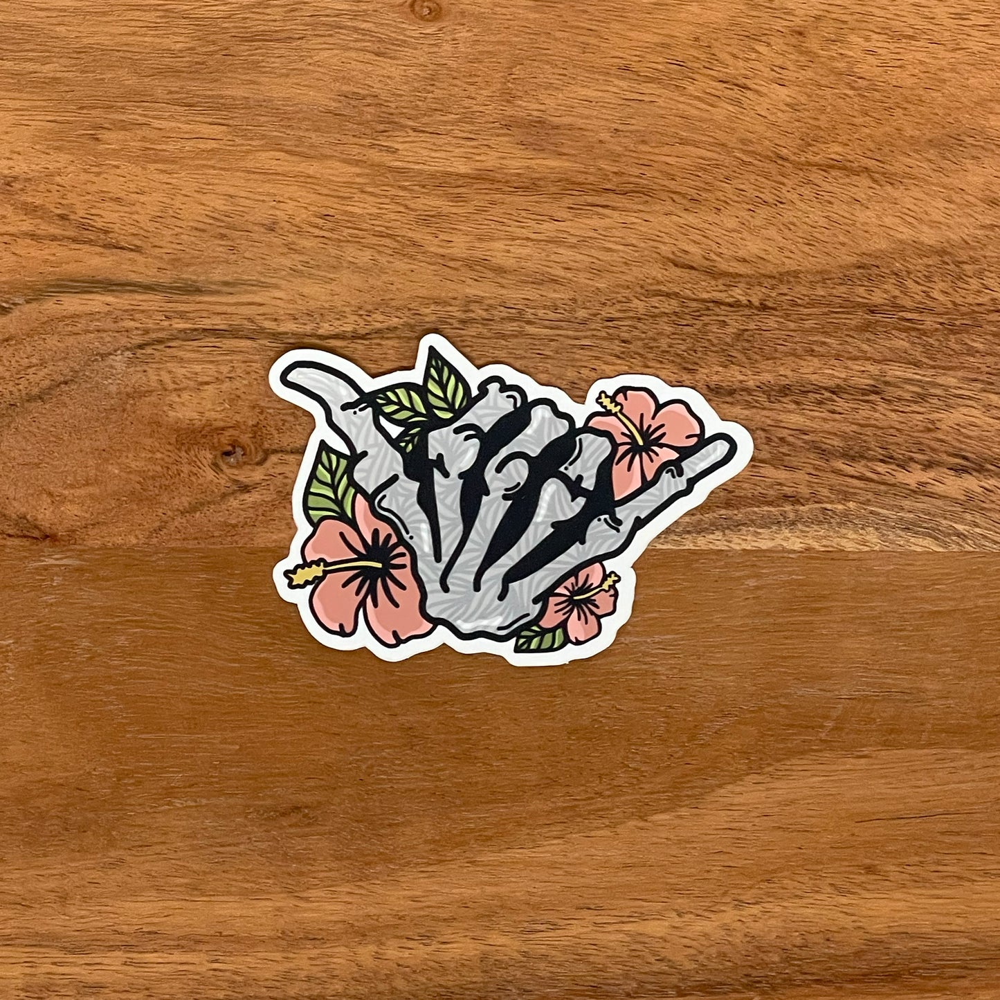A vinyl sticker featuring a hand-drawn illustration of a skeleton shaka with floral elements and a matte shimmer finish, placed on a wooden background.