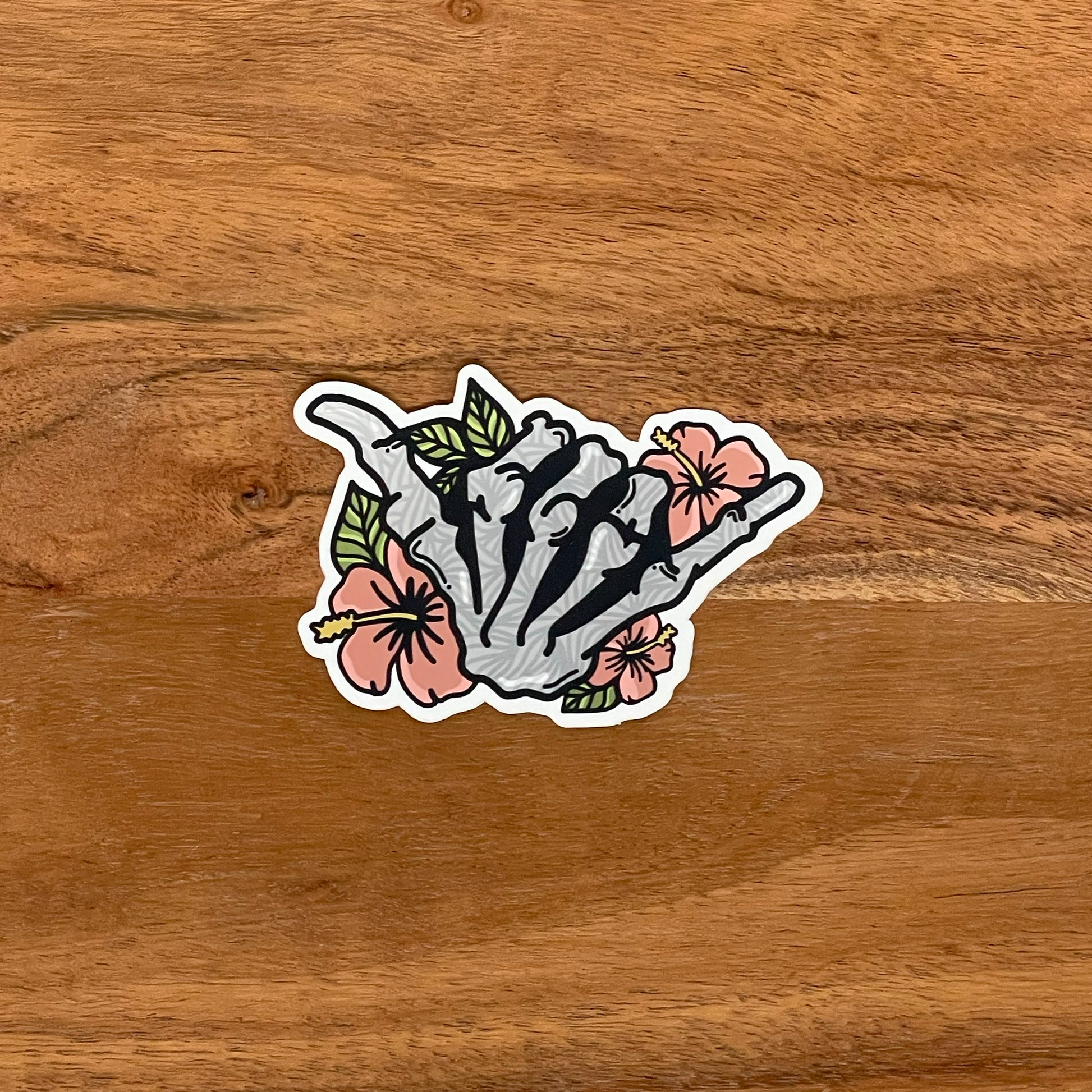 A vinyl sticker featuring a hand-drawn illustration of a skeleton shaka with floral elements and a matte shimmer finish, placed on a wooden background.