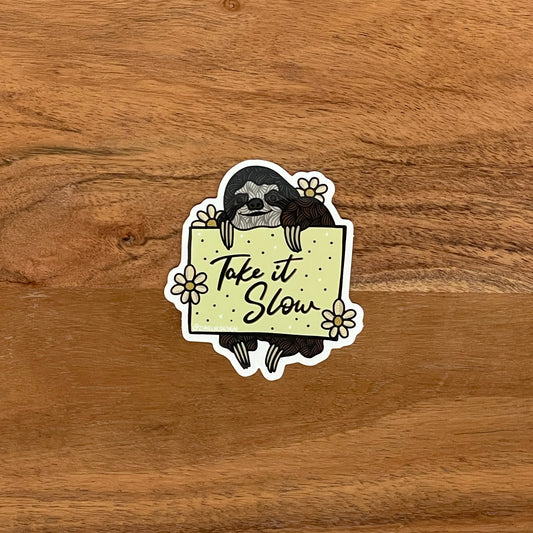 A vinyl sticker featuring a hand-drawn illustration of a sloth with a quote 'Take it slow', placed on a wooden surface.