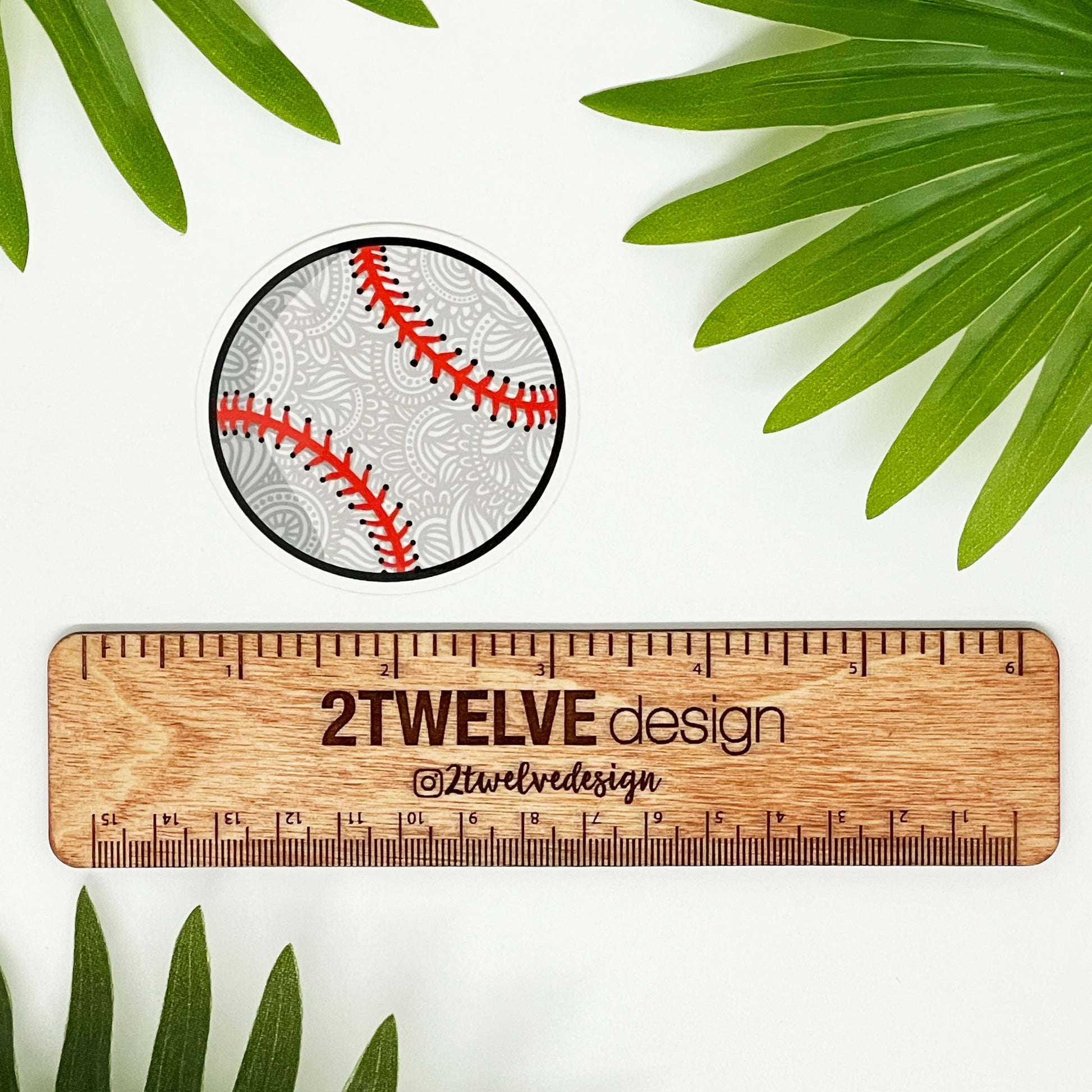 A small baseball sticker laying on a white background with surrounding leaves and a ruler to show actual size.