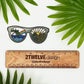 A sea to mountain, sunglasses illustration made into a vinyl die-cut sticker with clear back. Laying on a white background above a "2Twelve Design" ruler. Surrounded by palm leaves.
