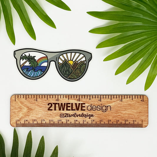 A sea to mountain, sunglasses illustration made into a vinyl die-cut sticker with clear back. Laying on a white background above a "2Twelve Design" ruler. Surrounded by palm leaves.