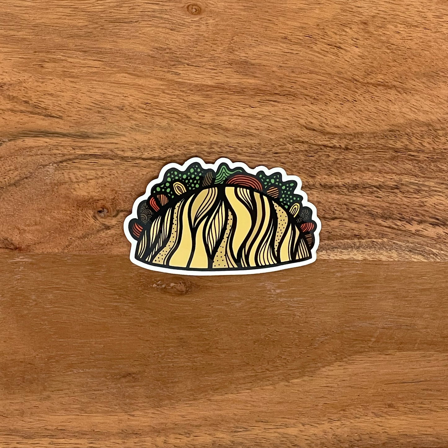 A fun, detailed illustrated taco sticker laying on a wood surface.