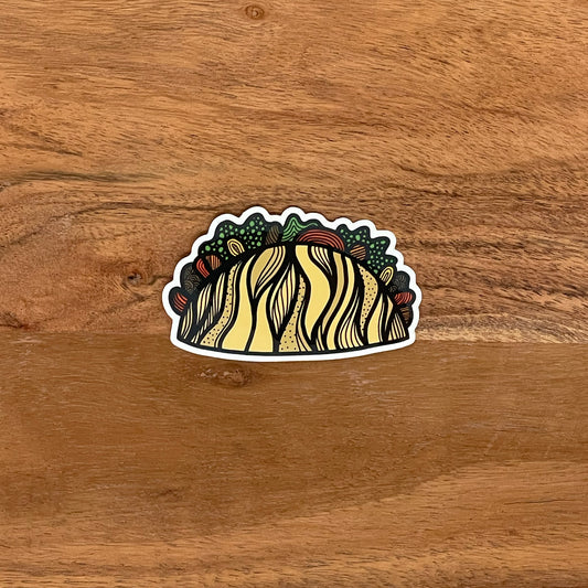 A fun, detailed illustrated taco sticker laying on a wood surface.