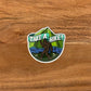 Bigfoot hiking in front of mountains and a river sticker. Laying flat on a wooden surface.