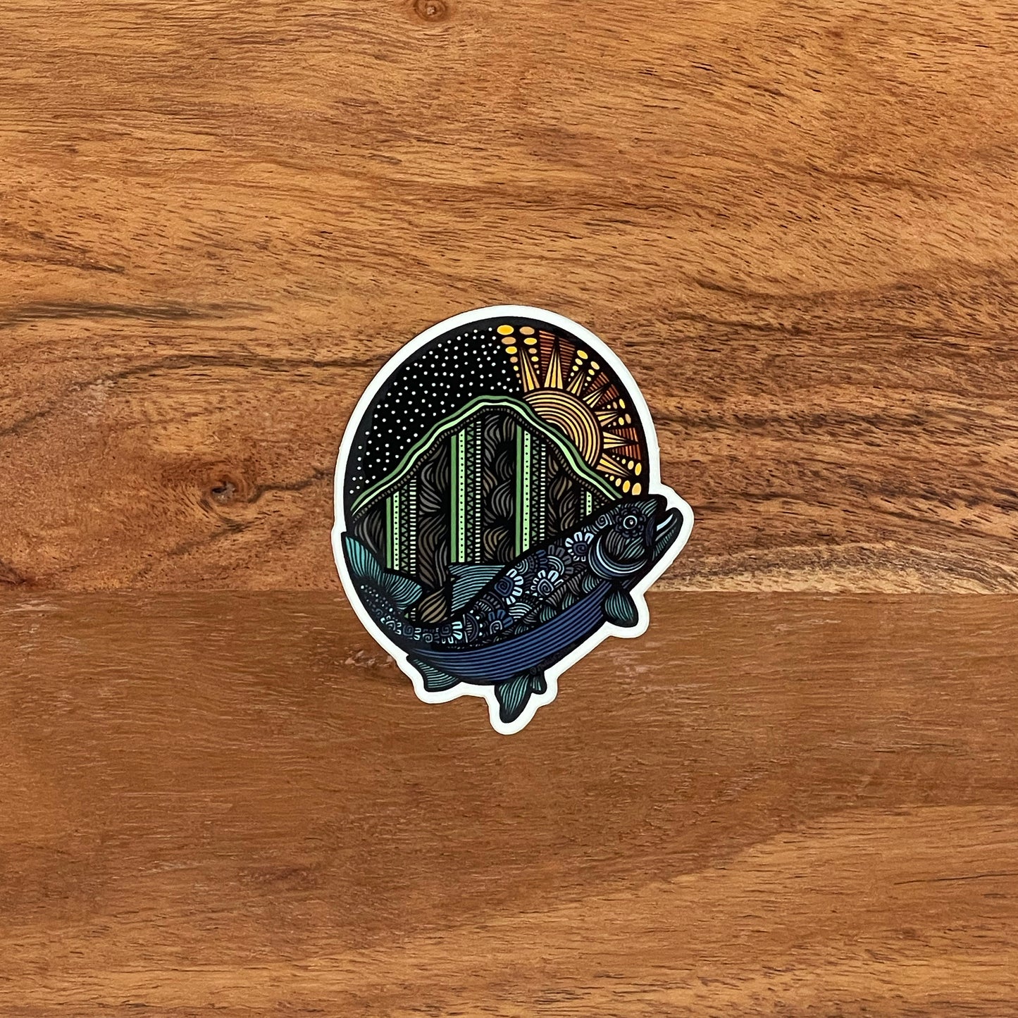 A detailed trout and mountain illustration made into a die-cut sticker. Laying on a wooden background.