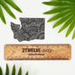 A sticker of Washington State with intricate hand drawn details. Laying on a white surface above a wooden "2Twelve Design" ruler surrounded by palm leaves.