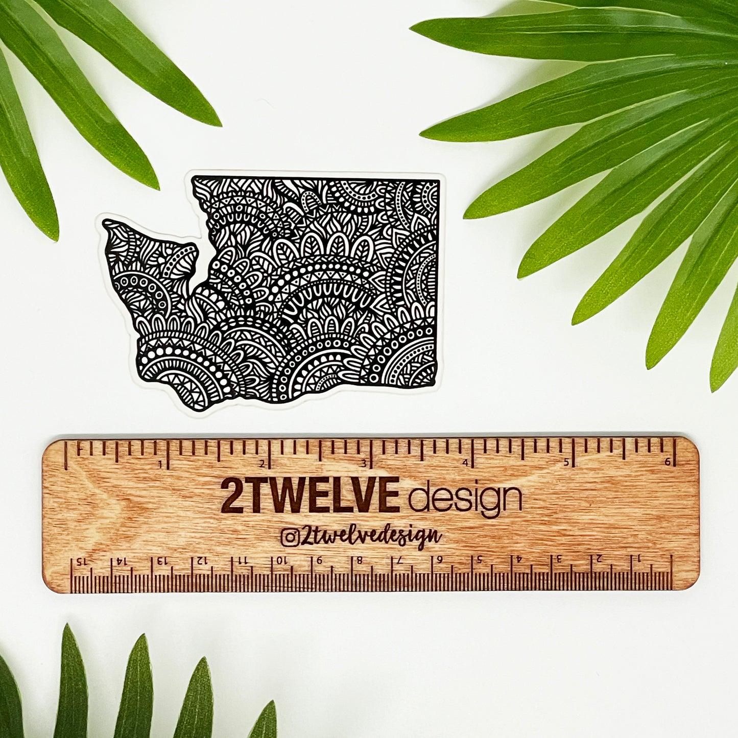 A sticker of Washington State with intricate hand drawn details. Laying on a white surface above a wooden "2Twelve Design" ruler surrounded by palm leaves.