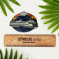 A diecut vinyl sticker of an illustrated ferry boat in Washington State. Laying on a white background above a wood "2Twelve Design" ruler, surrounded by palm leaves.