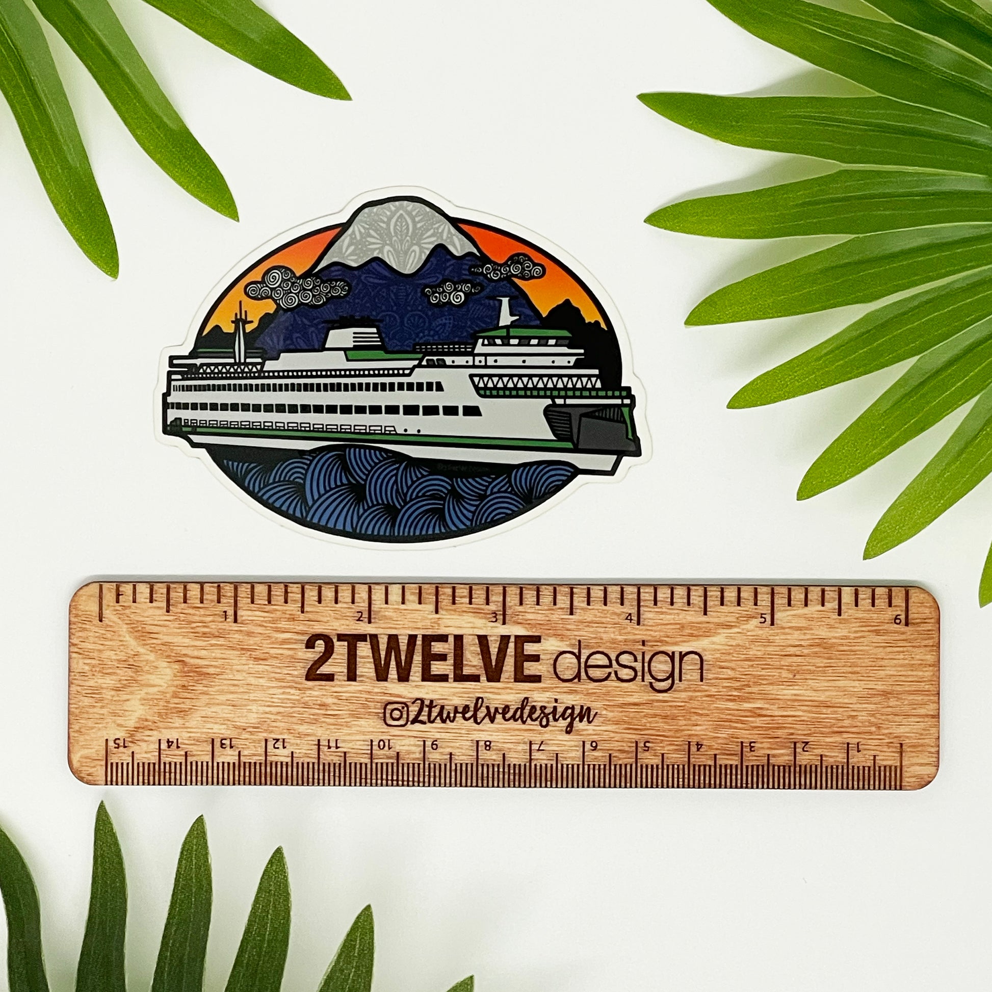 A diecut vinyl sticker of an illustrated ferry boat in Washington State. Laying on a white background above a wood "2Twelve Design" ruler, surrounded by palm leaves.