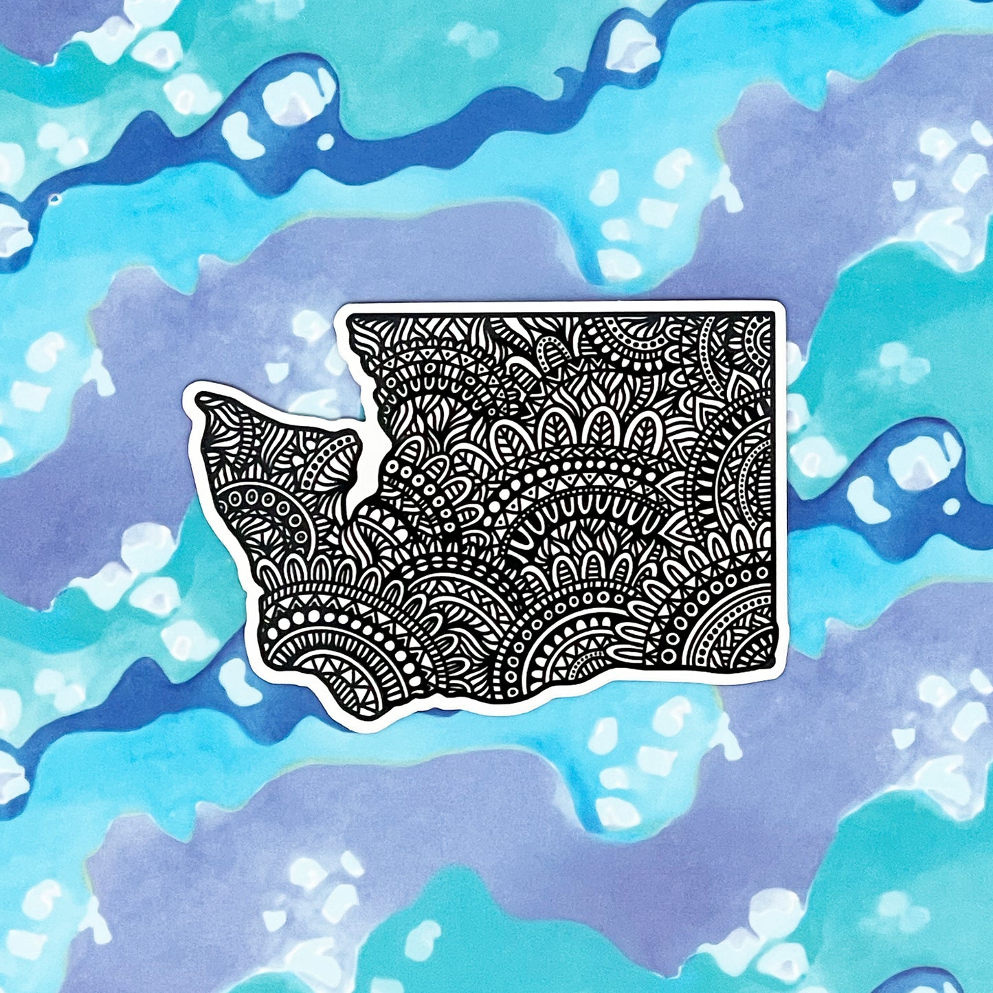 A sticker of Washington State with intricate designs filling the space. Laying on a blue and purple background.