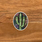 A cute, watercolor inspired cactus illustration made into an oval die-cut sticker laying on a wood surface.