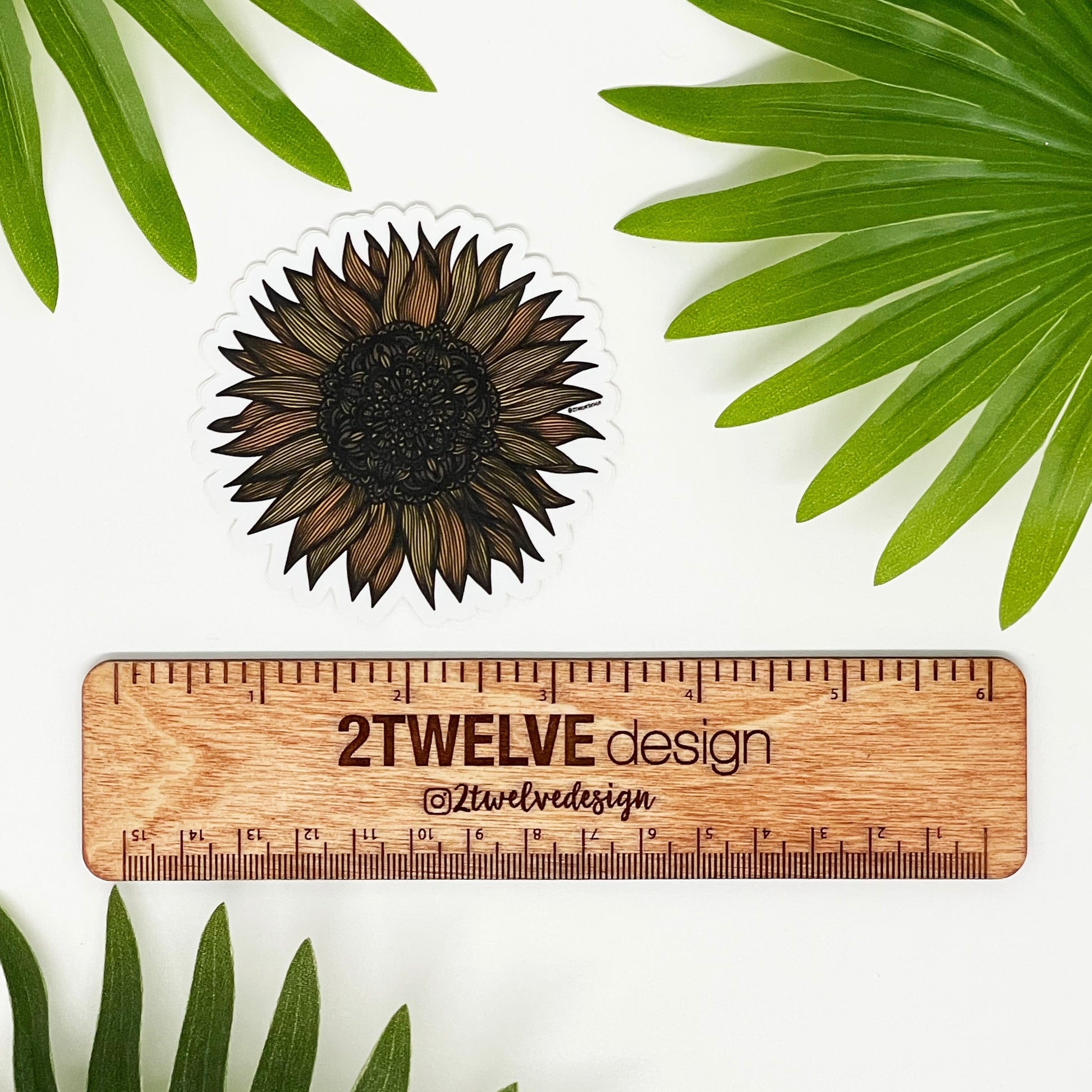 A zentangle inspired sunflower illustration made into a die-cut matte sticker. Laying on a white background above a "2Twelve Design" ruler, surrounded by palm leaves.